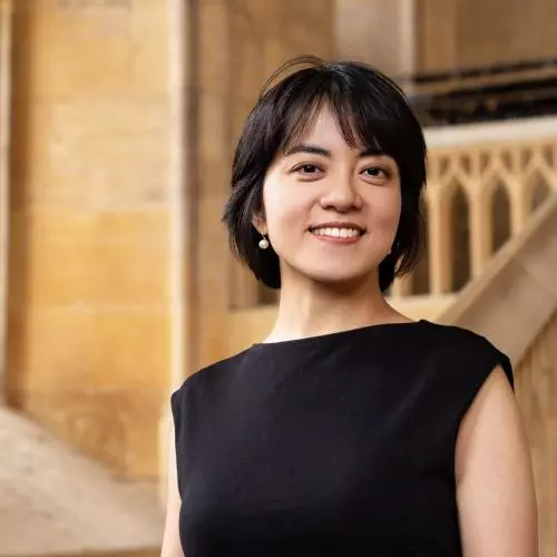 Yi Qie is wearing a black dress and pearl drop earrings with a short hair cut. She is smiling at the camera.