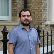 Dr Munoz has short black hair and black facial hair. He is wearing a short-sleeved blue shirt and standing outside a brick building. 